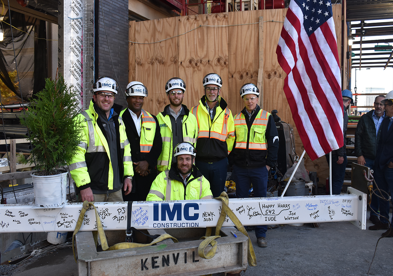 A group of IMC employees wearing safety vests and helmets smiling behind a steel beam with an IMC sticker and signatures.