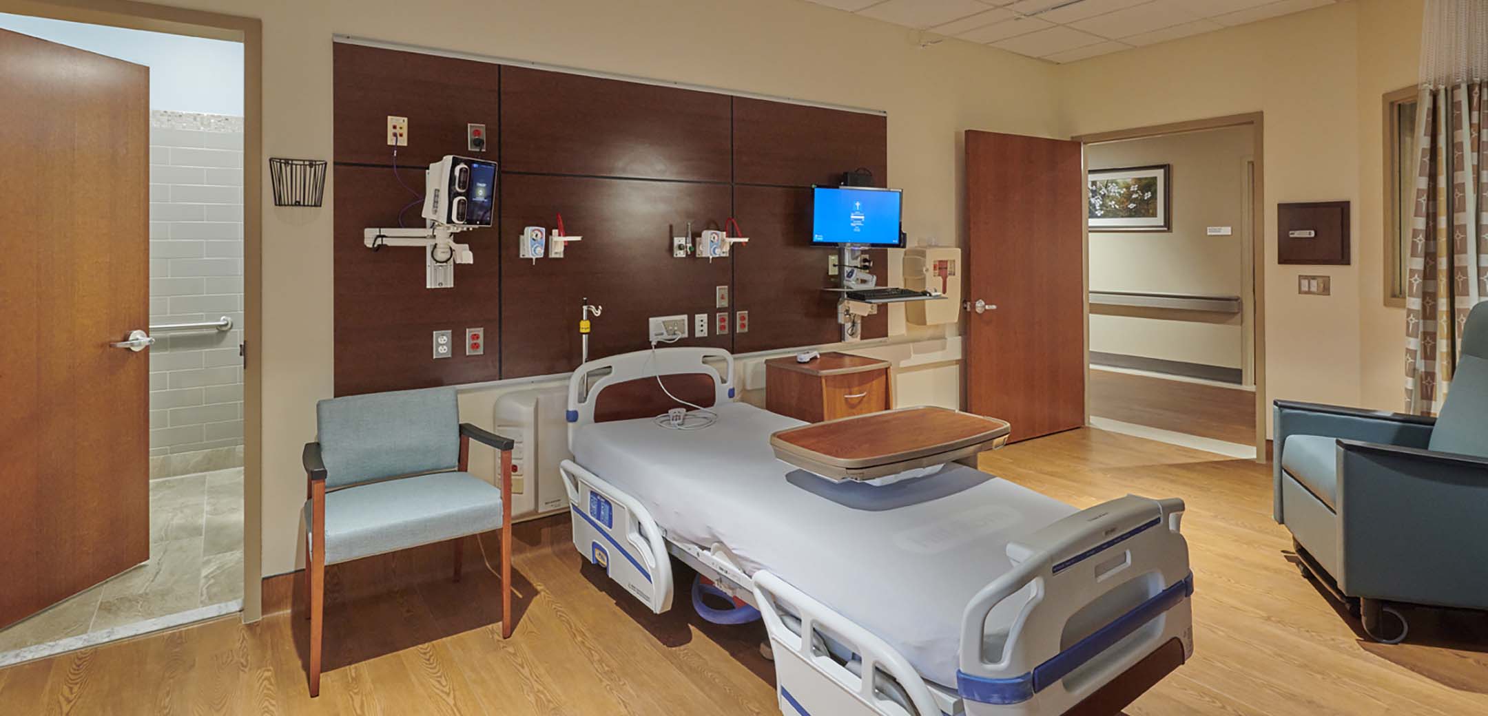 An interior view of the St Luke's Quarter building showcasing one of the patient rooms with seating chairs and patient bed with an accessible bathroom.
