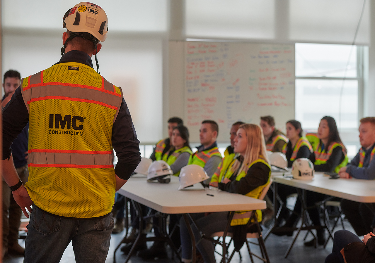 The up close view of the back of an IMC employee's safety vest with several IMC employees blurred in the background.