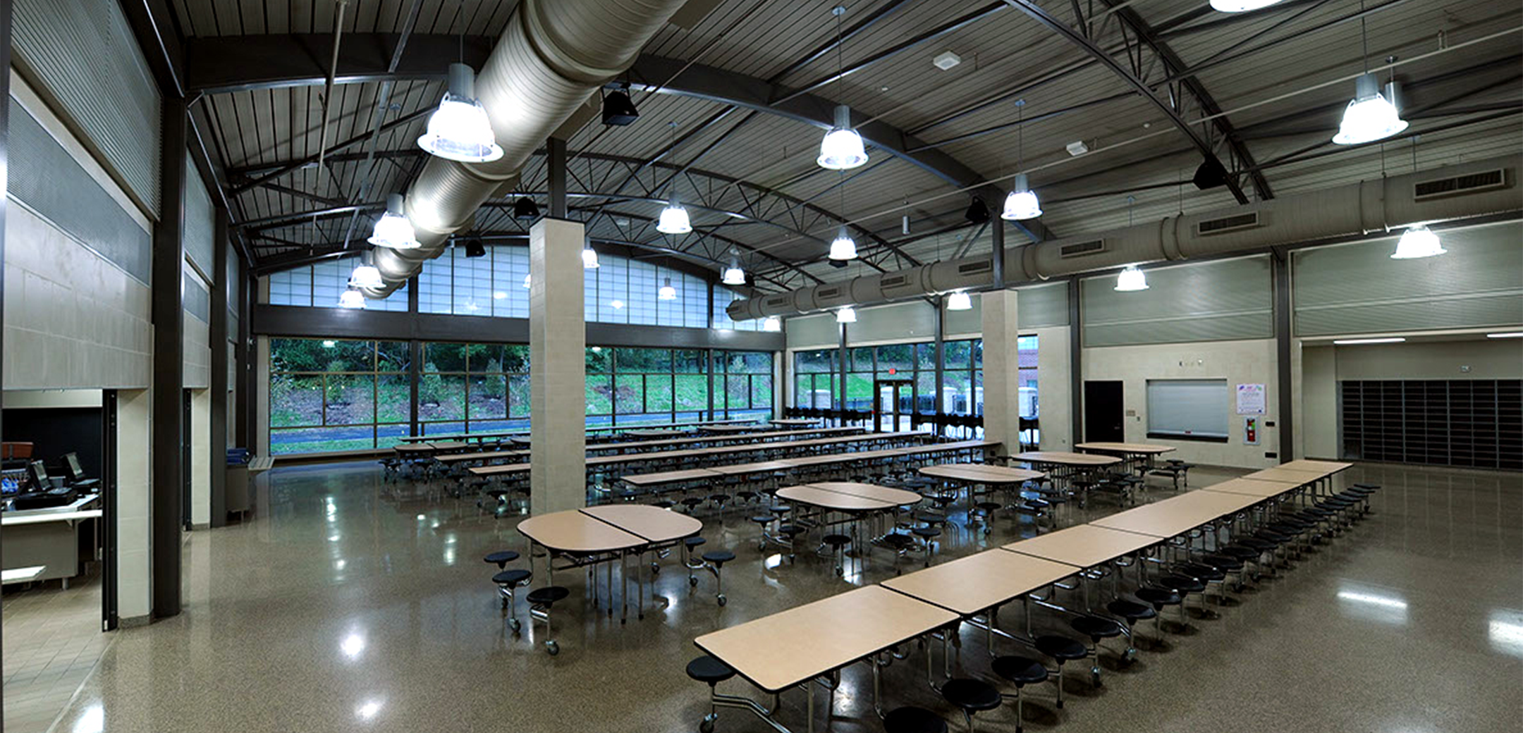 An interior view of the Phoenixville Middle School dining hall, showcasing the rows of dining seating, arched industrial design ceiling and all glass window back wall.