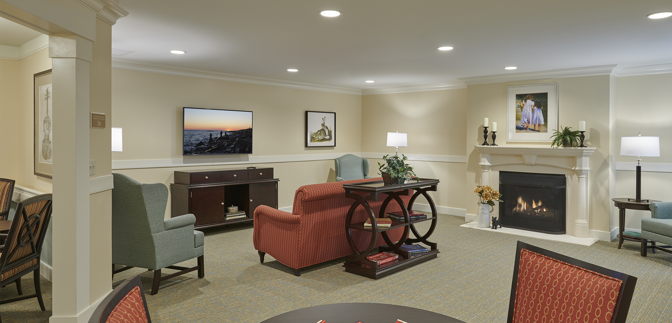 An interior view of the Artis Senior Living cream colored living room with a fireplace and couches towards the tv, table and seating in the foreground.