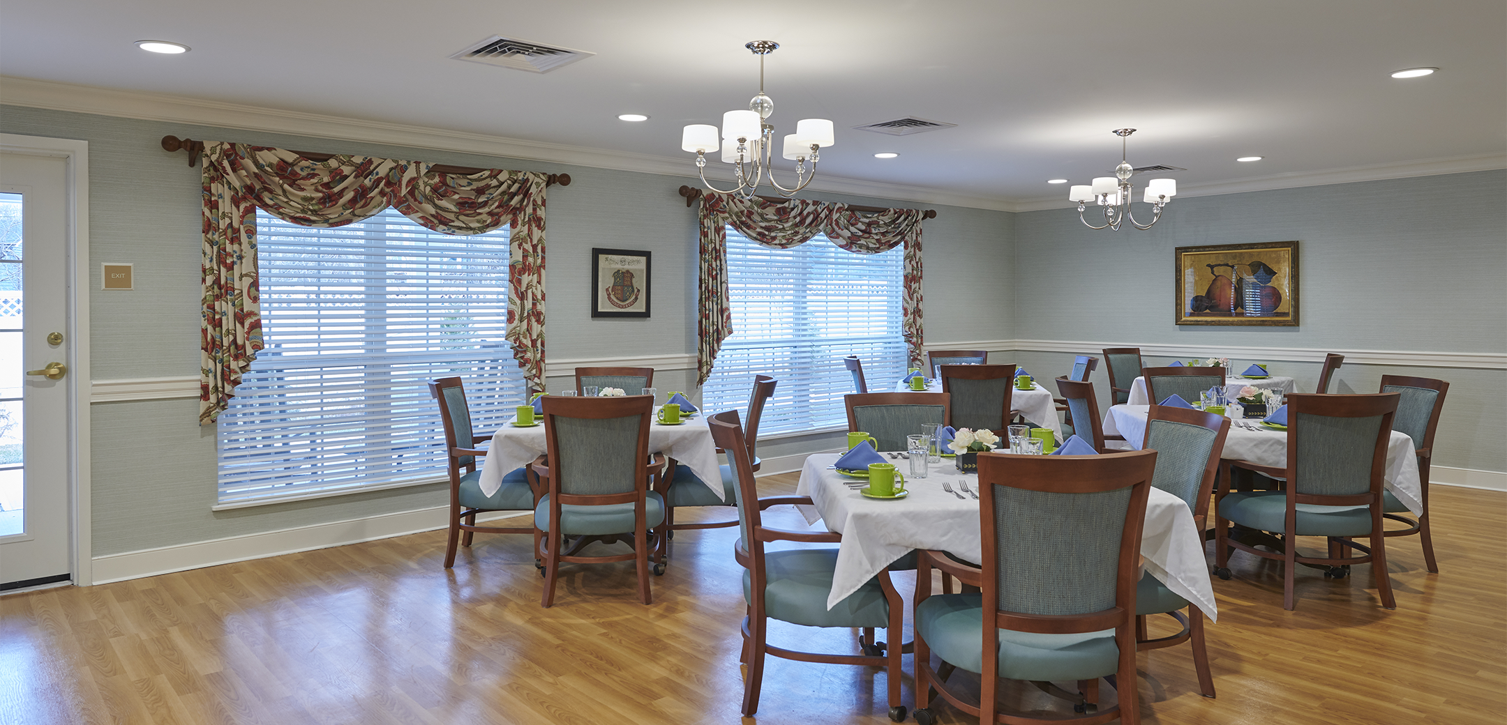 An interior view of the Artis Senior Living dining area with five tables and light blue cushioned chairs and wooden flooring.