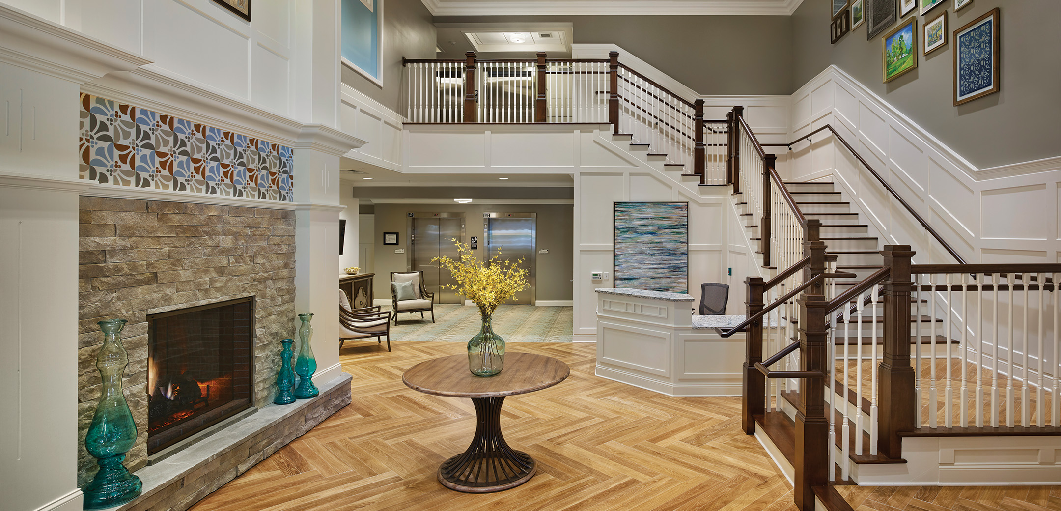 An interior view of the Arbor Terrace Morris Plains staircase leading up to the second floor with white accent panels and grey walls that have framed paintings on them and a brick fireplace on the right wall.