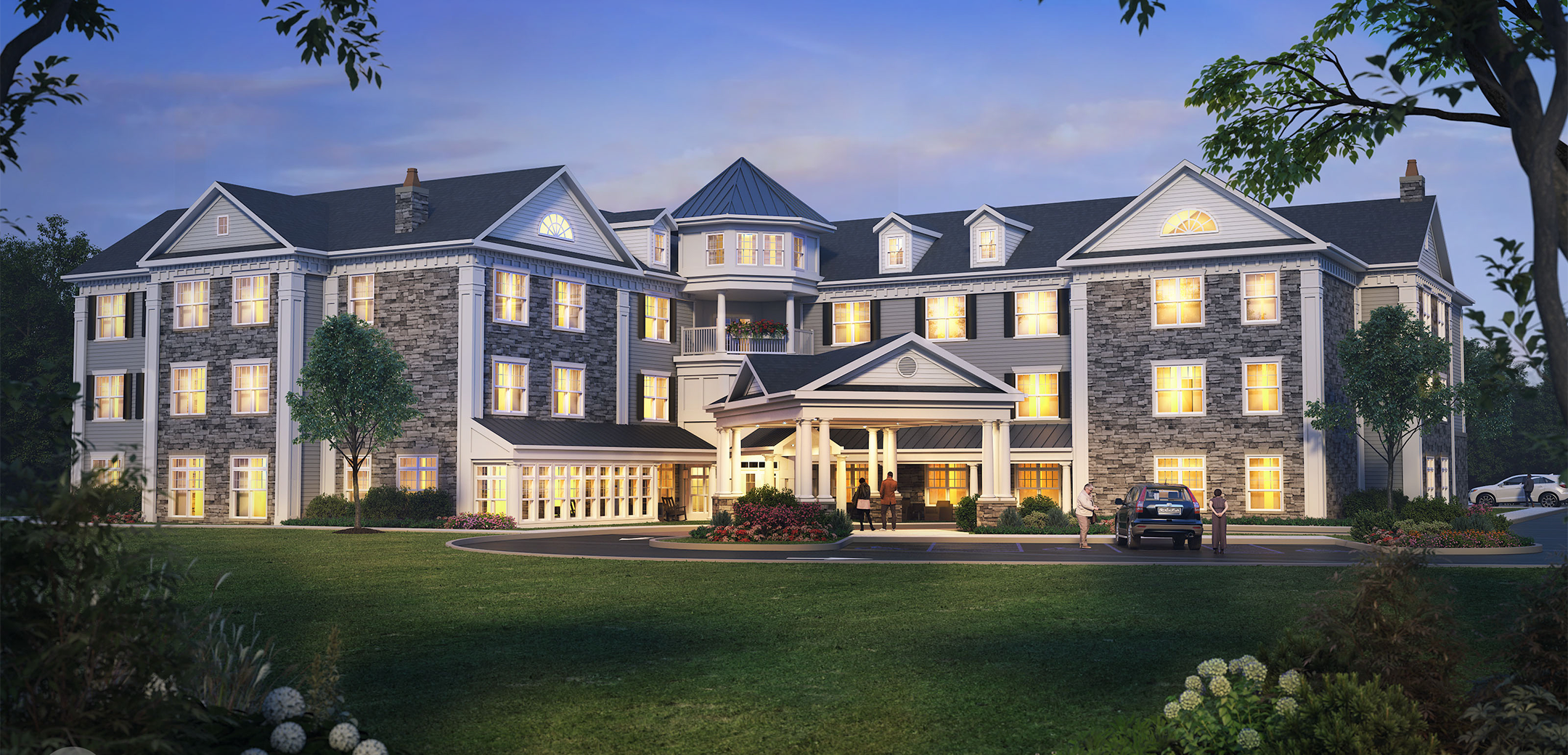 An exterior nighttime render of the Arbor Terrace Marlton stone brick, white wood accents and grey shingle roofing with a driveway, parking lot and grass lawn in the foreground.