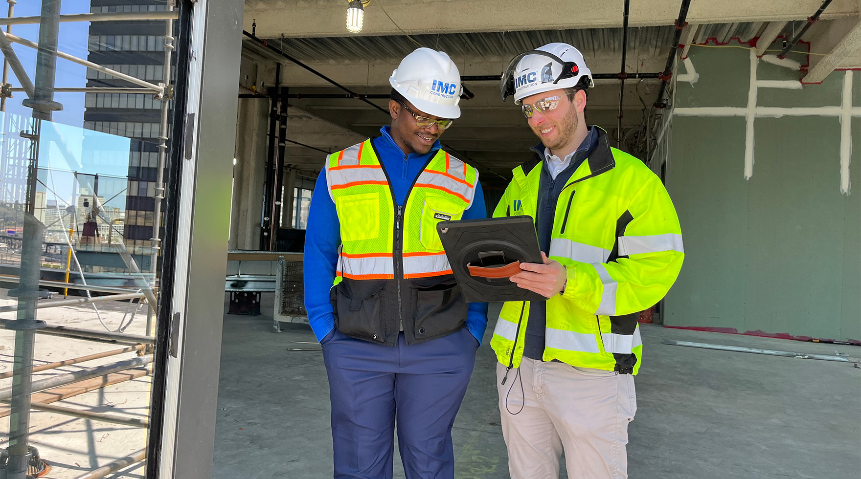 Two IMC employees looking at an IPAD on a construction site wearing safety helmets and vests.