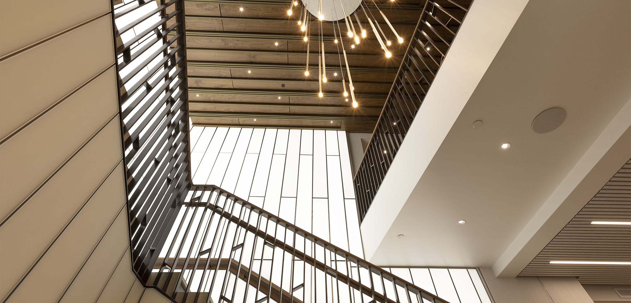 A bottom to top interior view of the Cohen Seglias staircase showcasing the light panel walls, brass colored metal ornate railings and a probe length hanging chandelier.