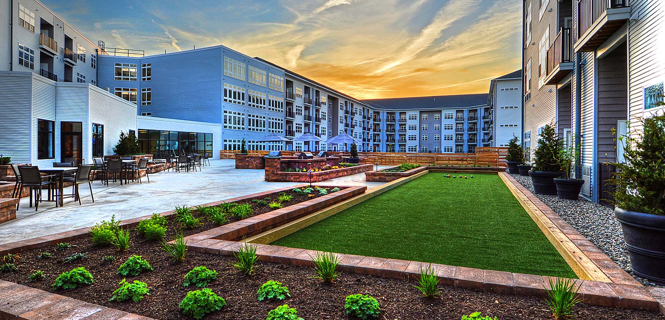 An exterior view of the Phoenix Village building showcasing the inner courtyard with a mini polo and outdoor table and chair sitting areas with the apartment windows in the background.