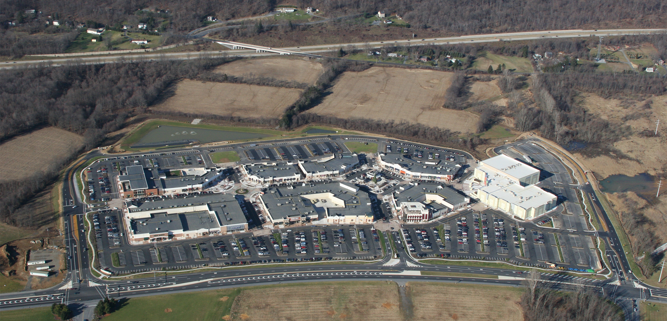 An aerial shot of the Promenade Saucon Valley building, showcasing the large layout with walkways inside, parking amendments surrounding the buildings, and forested general area.