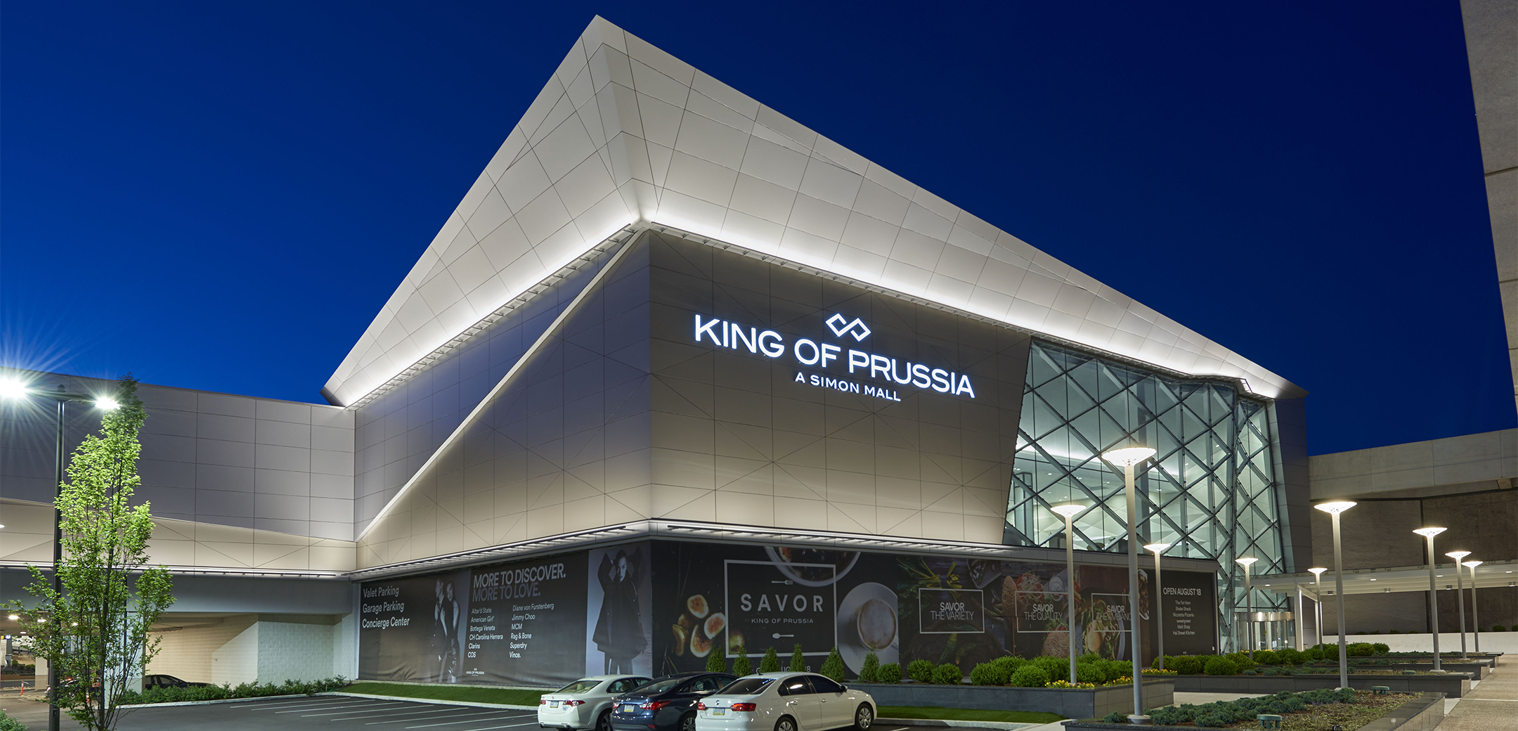 An exterior nighttime view of the KOP Expansion showcasing the angular modern design with lit strips and glass window accents, with a front parking lot.