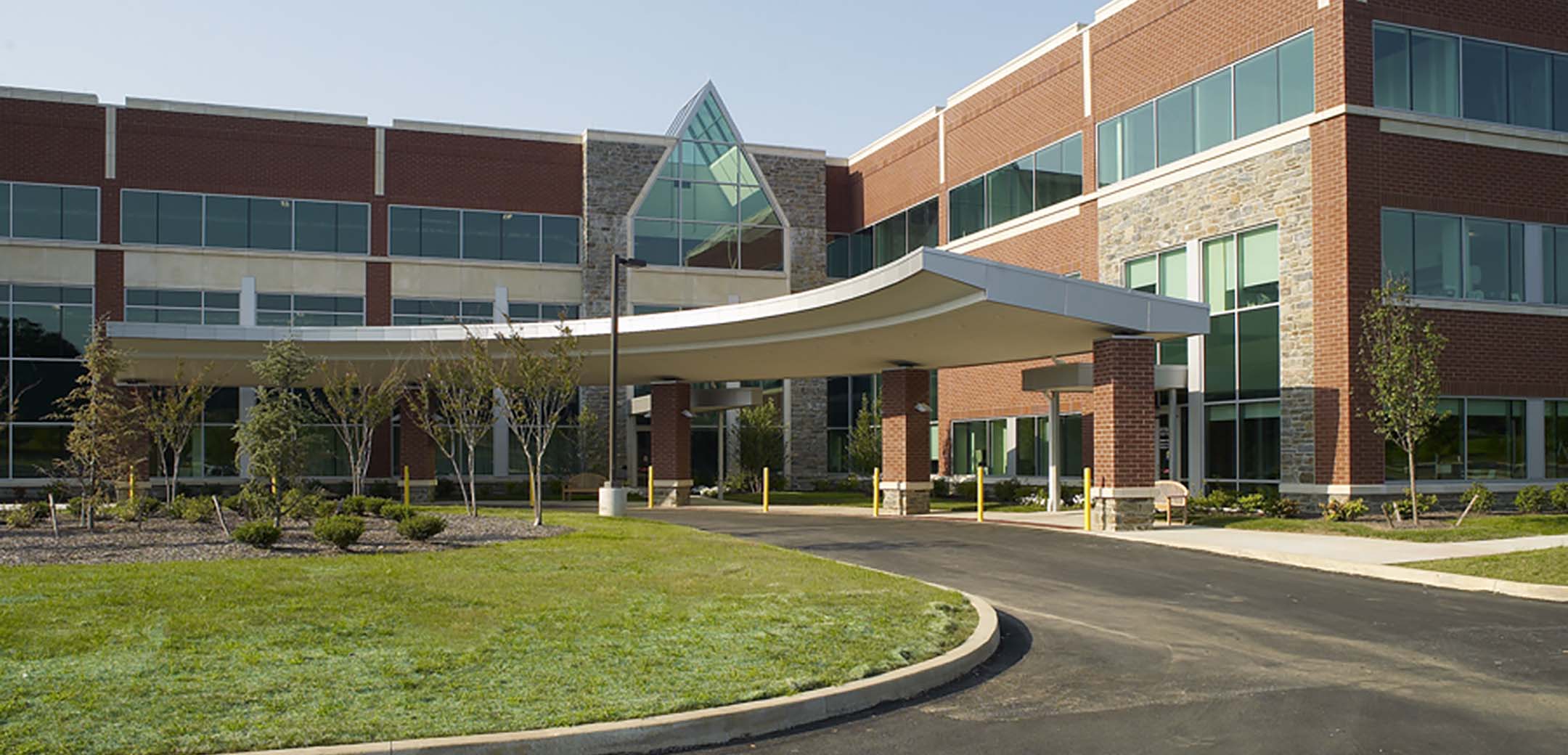 A view of the 3-story Main Line Health hospital from the front, showcasing the driveway leading up to the entrance and the grass lawn island Infront of it.