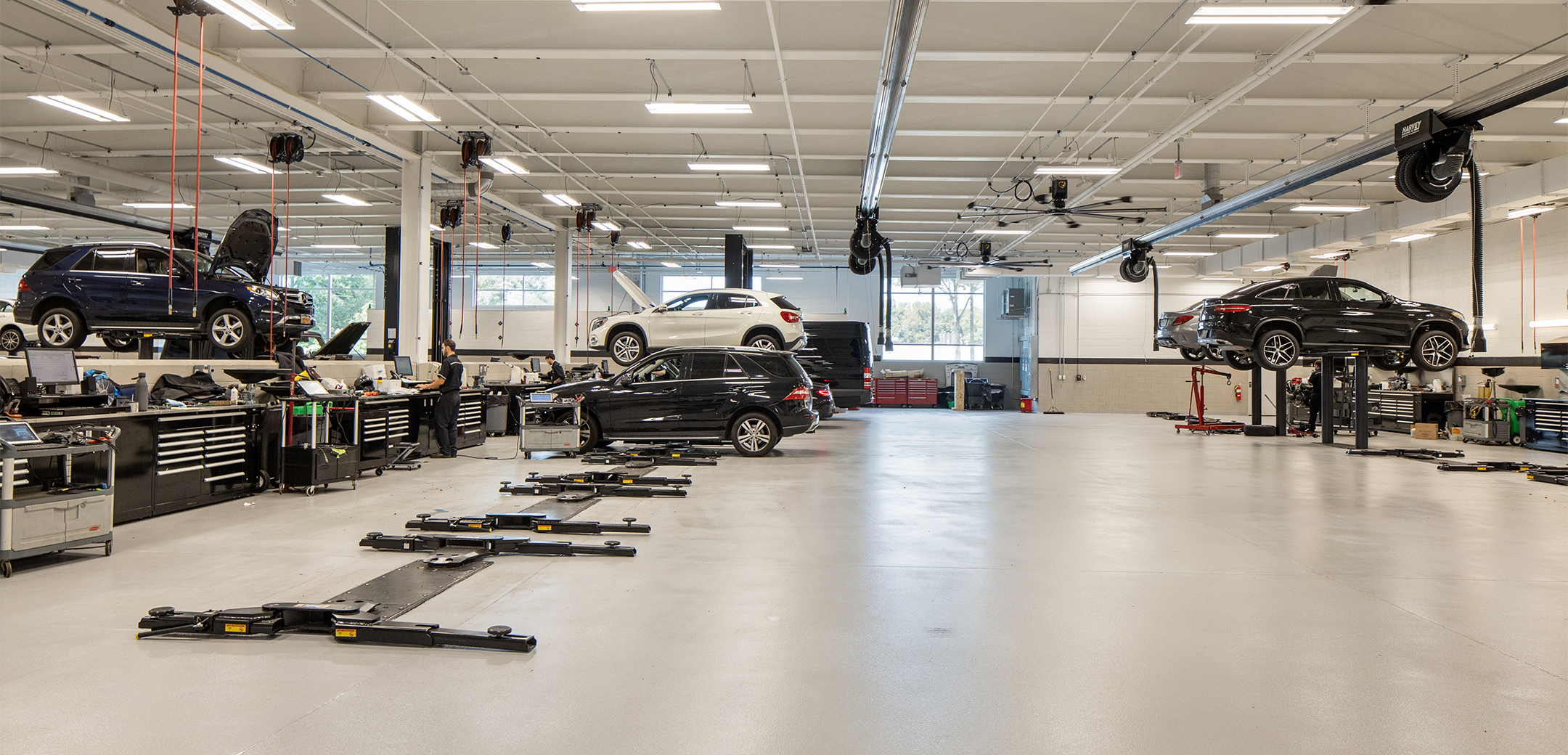 An interior view of Mercedes Benz of Caldwell garage showcasing the well lit tall ceiling, stationary car jacks, power jacks and cars in the background.