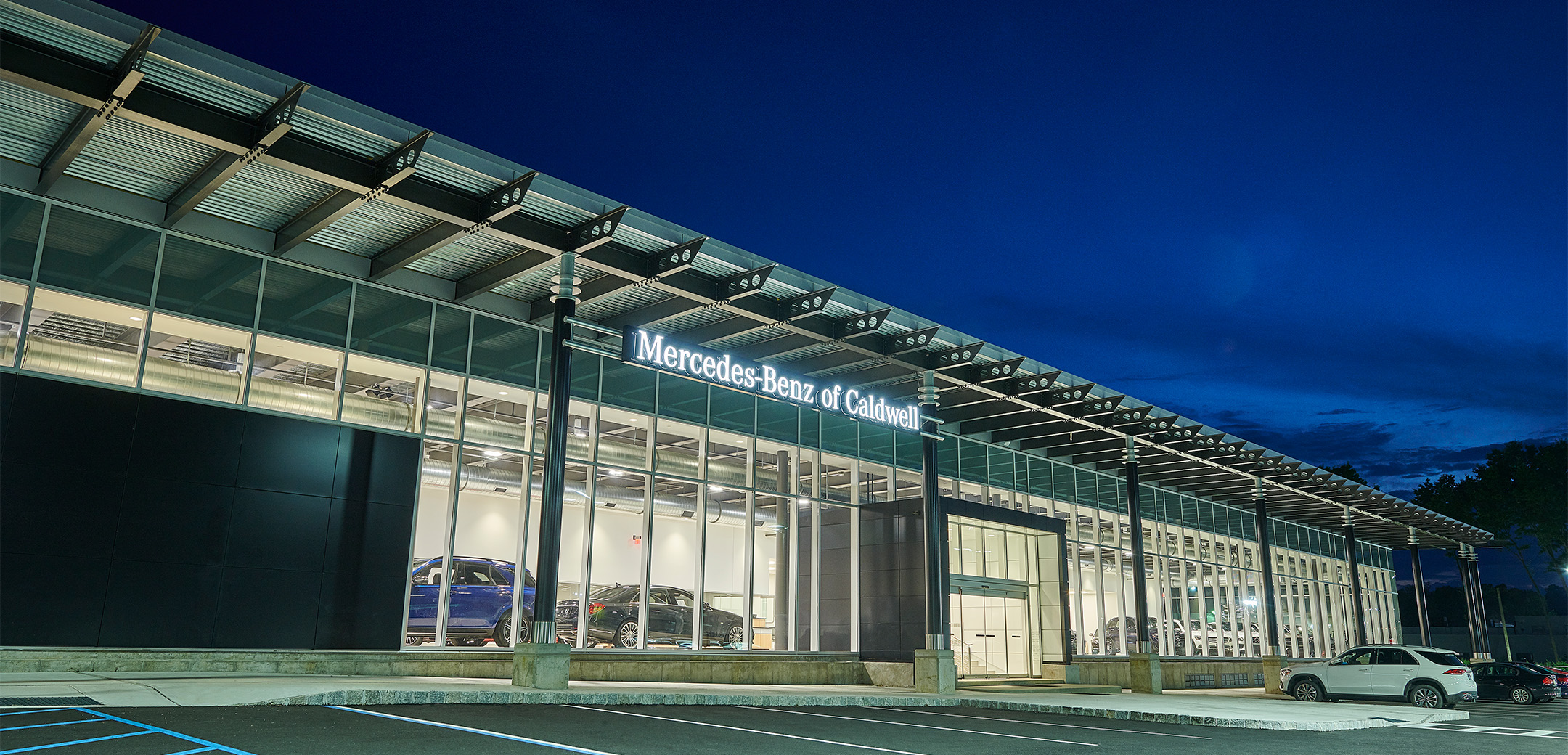 The exterior nighttime view of the Mercedes Benz of Caldwell single story building with an all glass panel car showcase wall, front entrance and main parking lot.