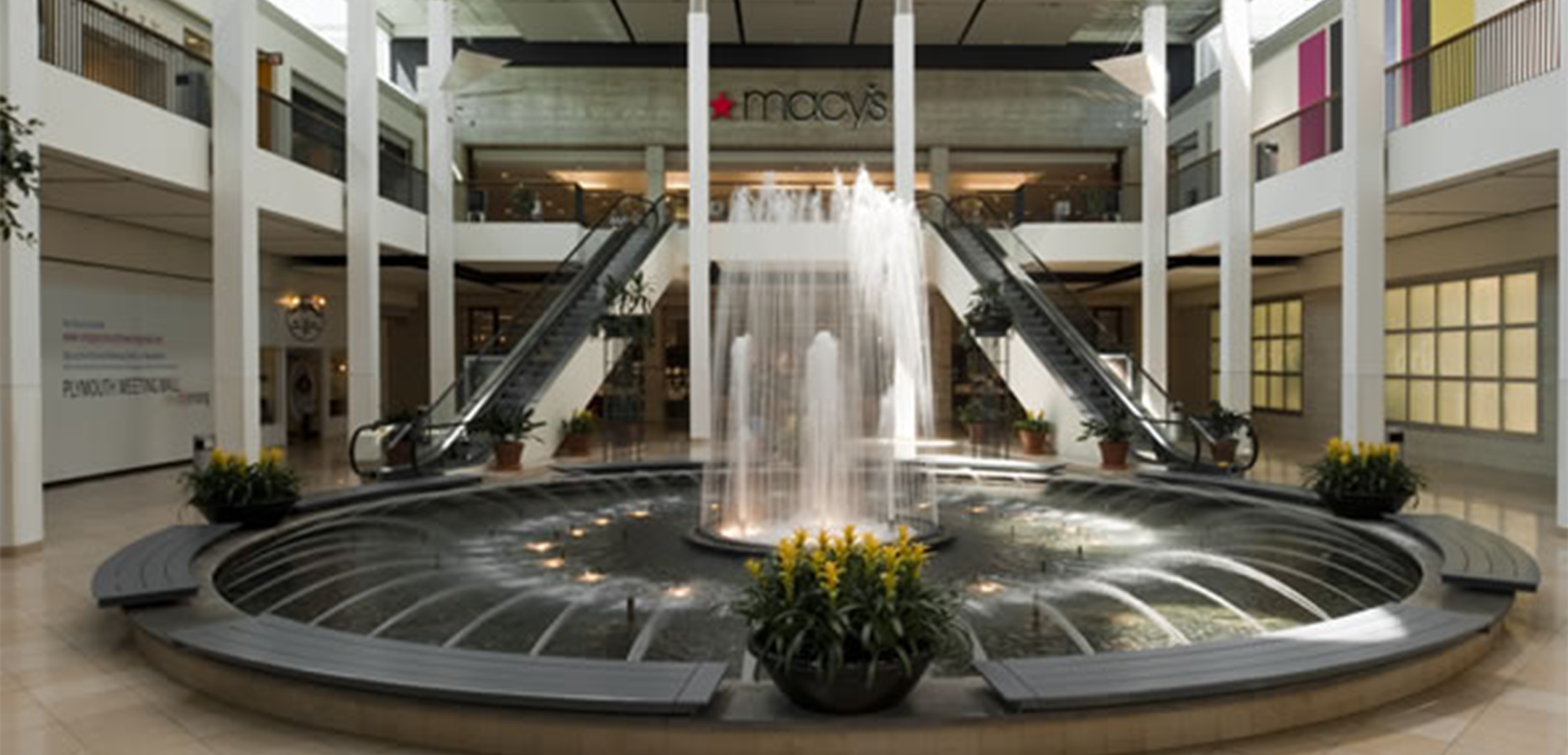 An interior close up view of the Plymouth Meeting Mall circular fountain with benches and flower pots on the edges and escalators in the background going up to the second floor.