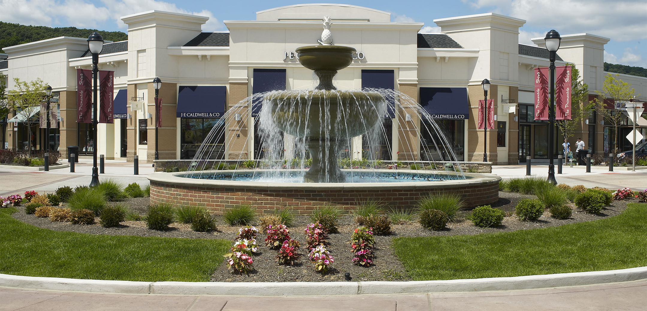 A close up exterior view of the Promenade Saucon Valley main square fountain with a grass and bush landscape encircling it with the building in the background.