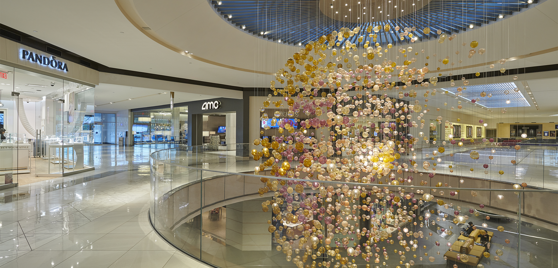 An interior view of the Shops at Riverside mall, showcasing the circular second floor balcony hole peering down to the first floor and a large light installation made of small yellow bubbles going down the balcony.