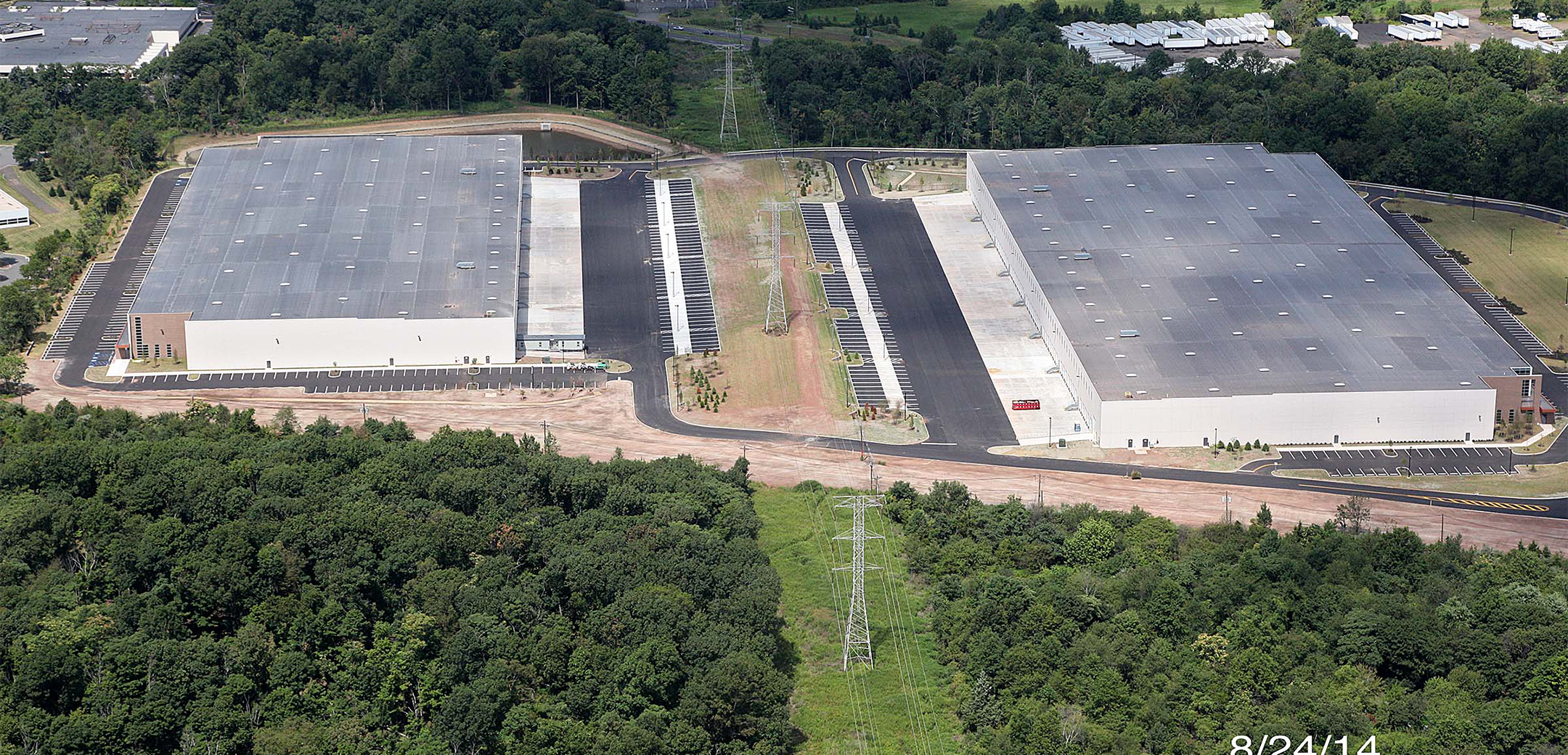An aerial view of the South Washington Park buildings showcasing the two warehouses, parking lots, overall layout power lines going down the center and a green forest in the foreground.