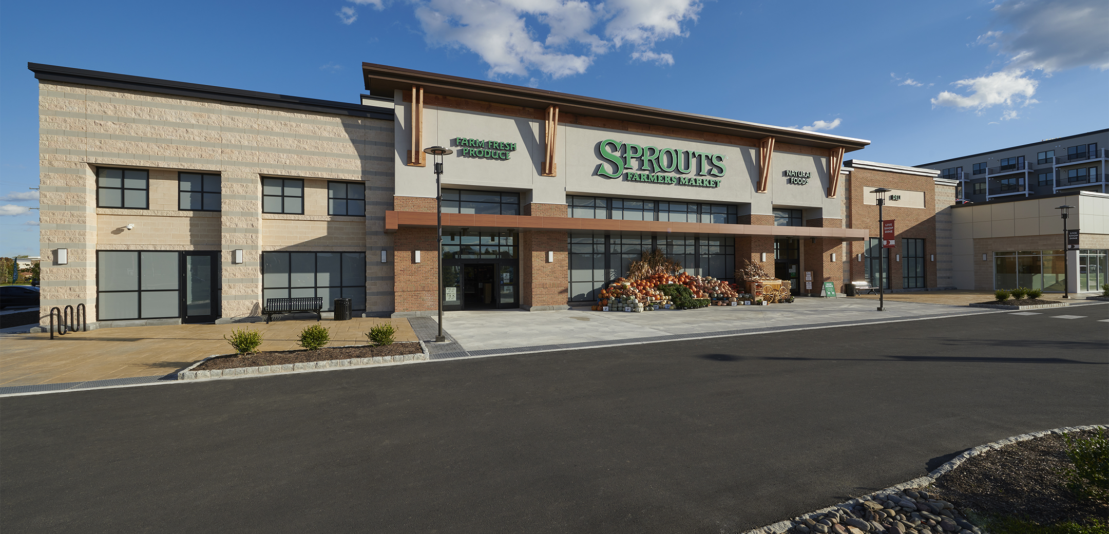 The exterior of the Sprouts Farmers Market brick building showcasing the front entrance, driveway, storefront flowers and a road in the foreground.