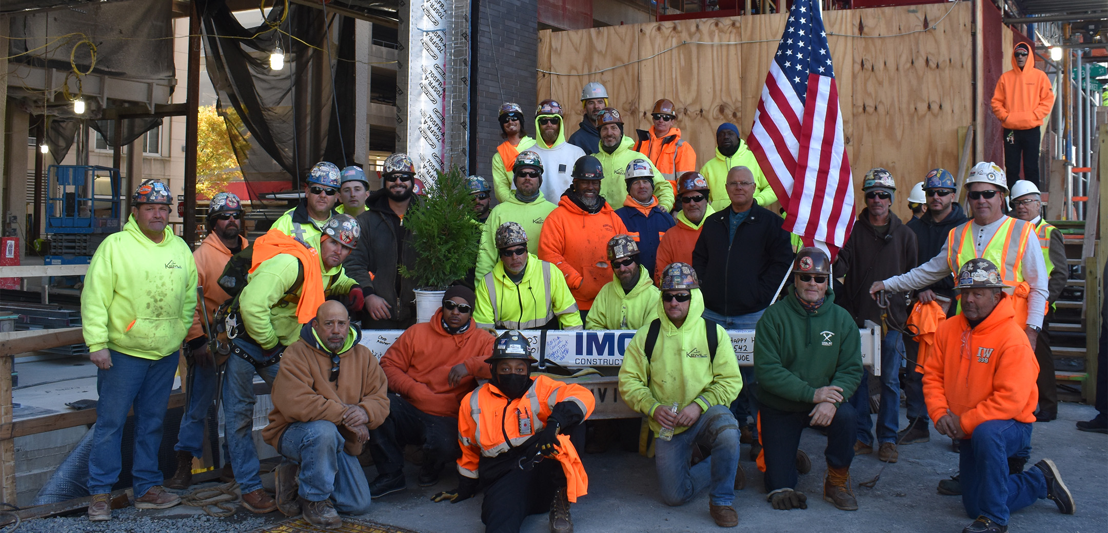 A group of construction workers wearing safety helmets and vests in front of the topping out beam on the 2222 Market Street project site.