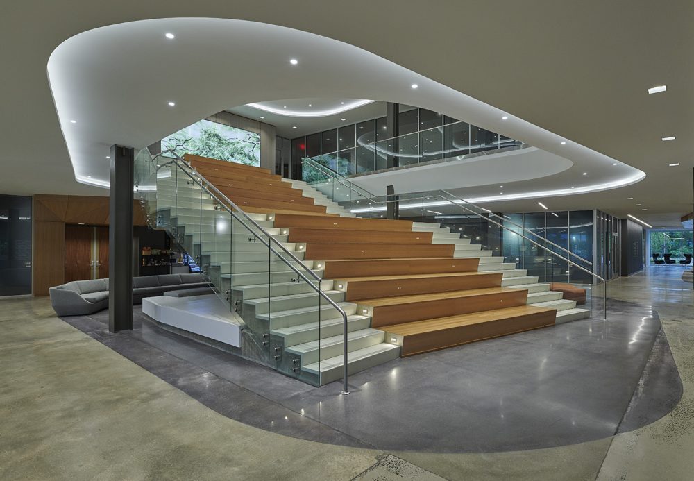 The larger interior staircase of the Blue Bell office building with wood and cement stairs accompanied with glass guardrails.