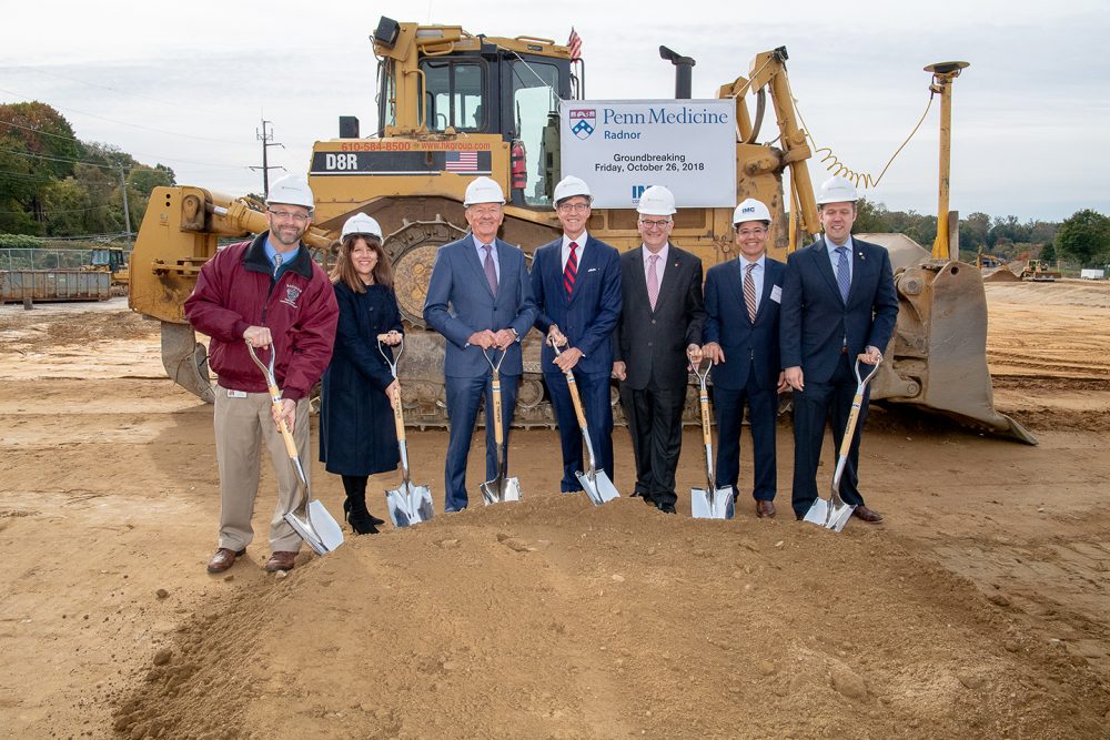 A group of project members with IMC Construction hardhats and shovels holding shovels for the ground breaking in front of heavy machinery with a sign for Penn Medicine Radnor behind them.