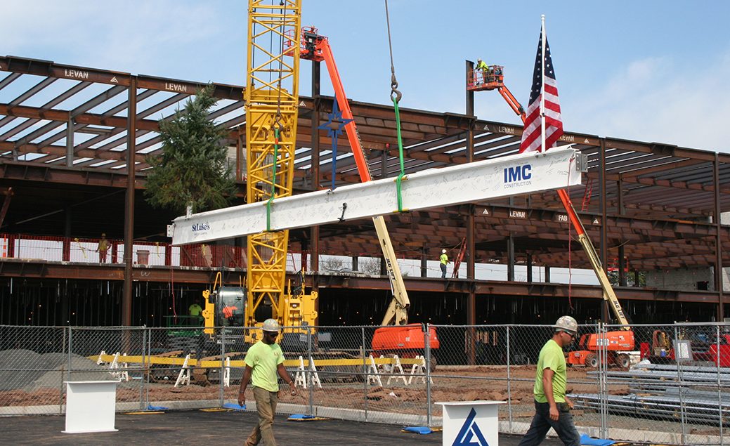 A large white, iron beam being lifted by crane to the top of the new building with an American Flag attached and two IMC construction workers below.
