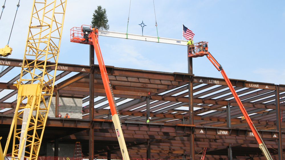Two cranes lifting a white, iron beam to the top of a building with a tree and an American flag attached to the beam.