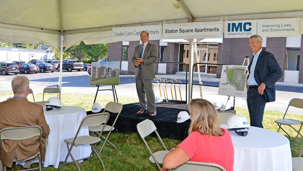 A man in a gray suit standing on a small station in front of three other people speaking at the groundbreaking of the Station Square Apartments project.