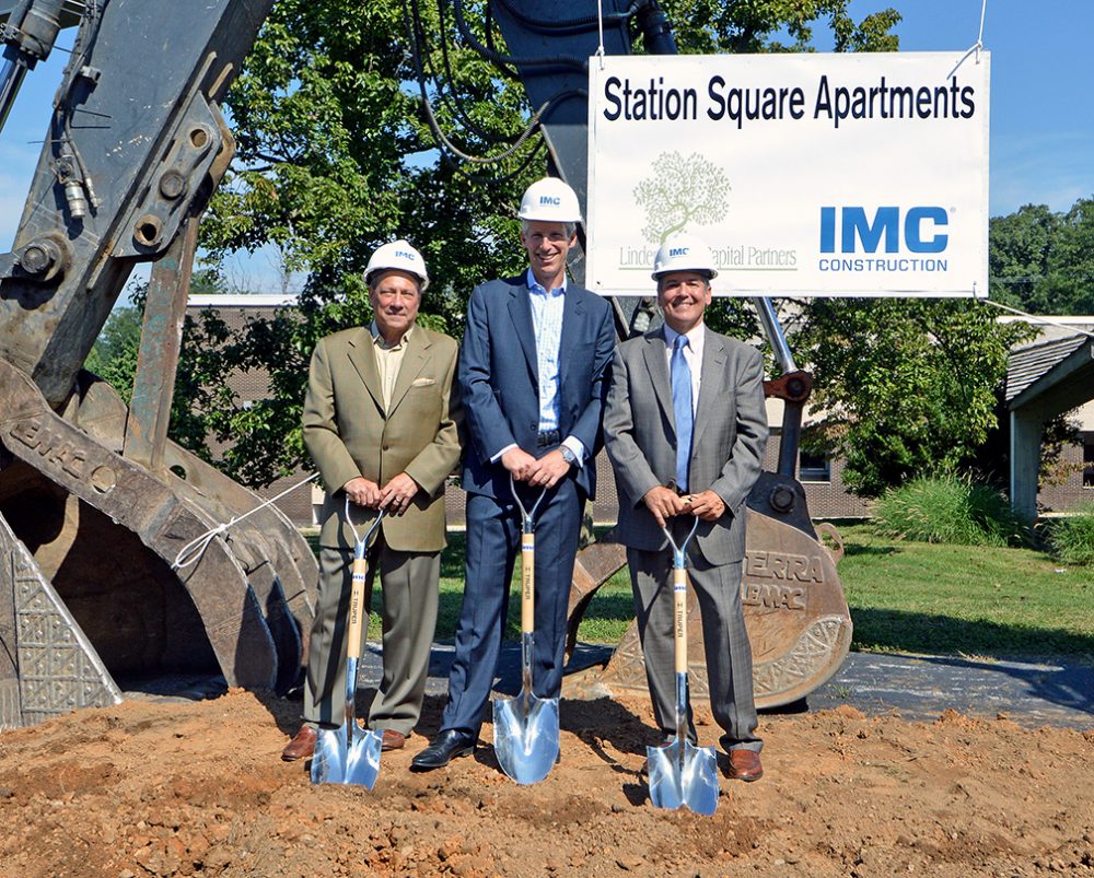 Three IMC Construction employees wearing IMC hardhats and holding shovels in front of heavy machinery with a sign that says "Station Square Apartments" behind them.