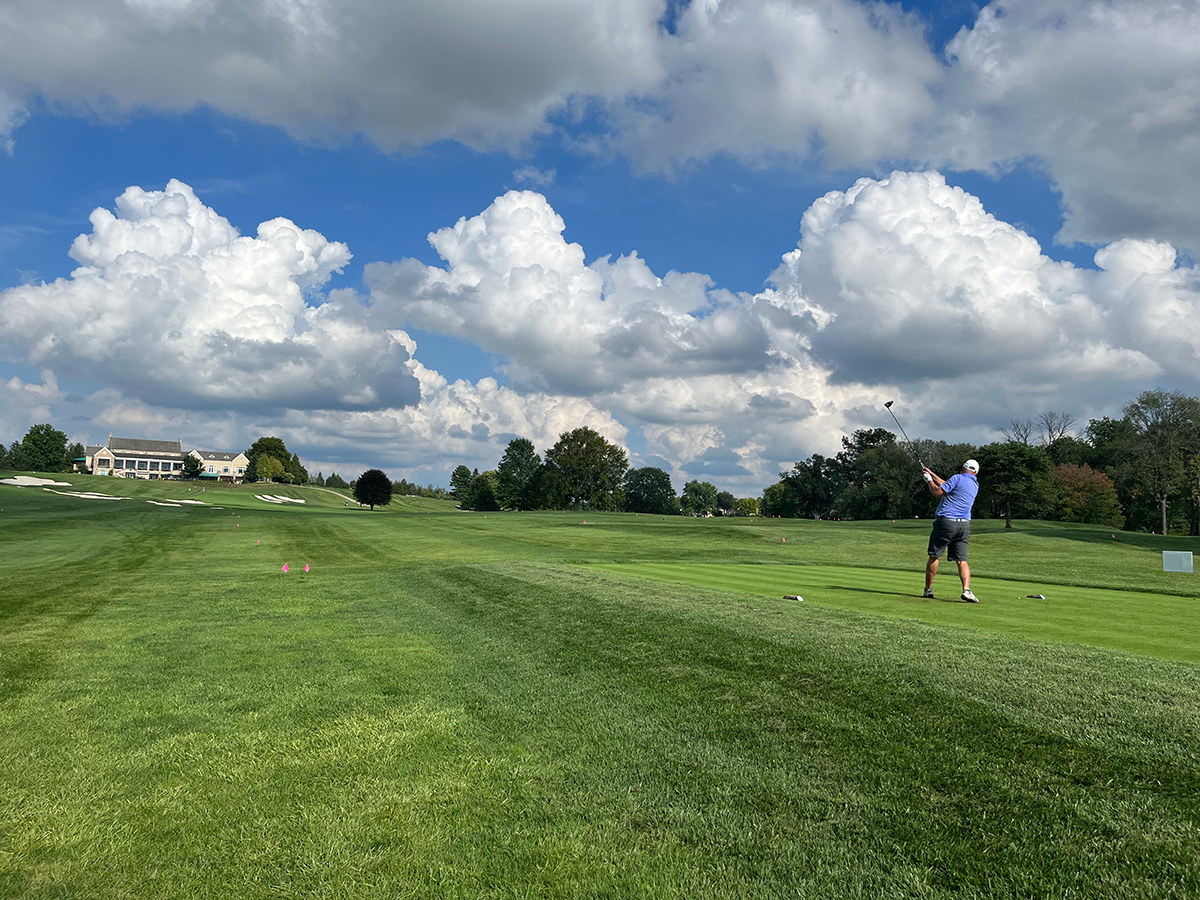 A golfer hitting a ball on a green golf course with blue skies and clouds